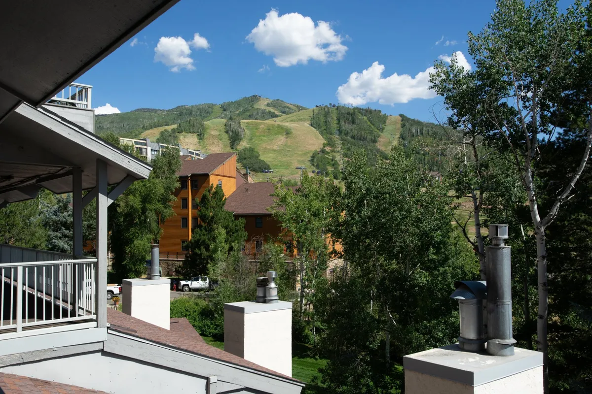 The image showcases a tranquil balcony with seating, overlooking lush green trees and majestic mountain views. The building's mountain-inspired architecture is visible, and the scene is bathed in soft sunrise light, suggesting peaceful moments spent outdoors. The setting evokes relaxation and connection with nature, ideal for both unwinding and enjoying outdoor activities like hiking or photography.