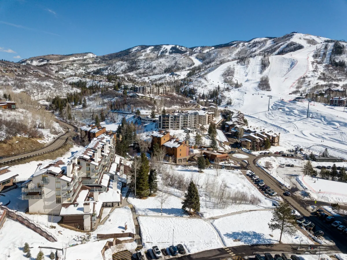 This image features snow-covered mountains in the background, ski slopes ready for winter adventure, and residential buildings that establish the proximity to both the slopes and amenities. A visible parking area hints at easy access and convenience for visitors with vehicles, including those with EVs.
