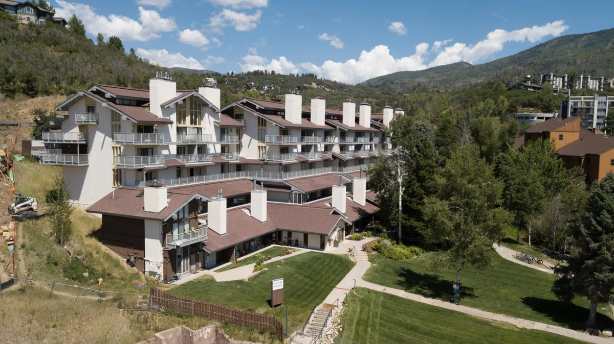 A tranquil view of a multi-story building set against a landscaped lawn and majestic mountain backdrop. The scene includes wildflowers and hints of outdoor leisure, with paths that invite exploration and scenic relaxation.