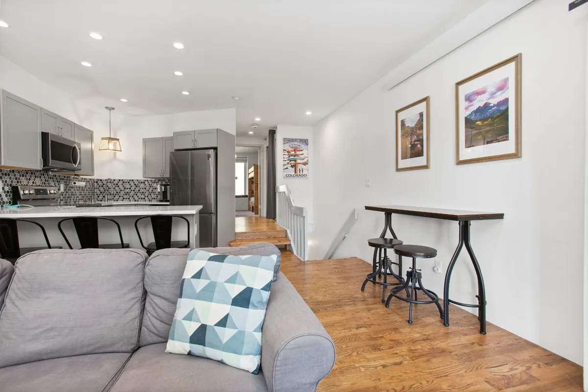 The image shows an open-concept living and dining area. A comfortable sofa with geometric pillows faces a kitchen island with sleek bar stools. The space is well-lit and social, with artwork on the wall and a layout that allows for easy flow between relaxing, dining, and cooking.