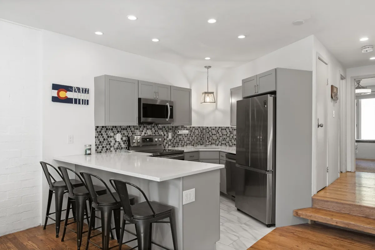 A modern kitchen is showcased here, featuring a central island, stainless steel appliances, black bar stools, and a decorative backsplash. The space looks newly renovated with gleaming counters and ample seating, ideal for group meal prep and casual gatherings.