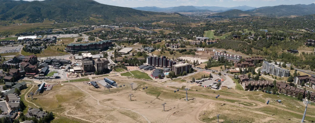 This image captures the lively base area with mountain views, ski lifts, residential buildings, and a bustling commercial strip. The scene evokes the excitement of an alpine village, with visible access to local shops, dining, and year-round activities for families to explore on foot.