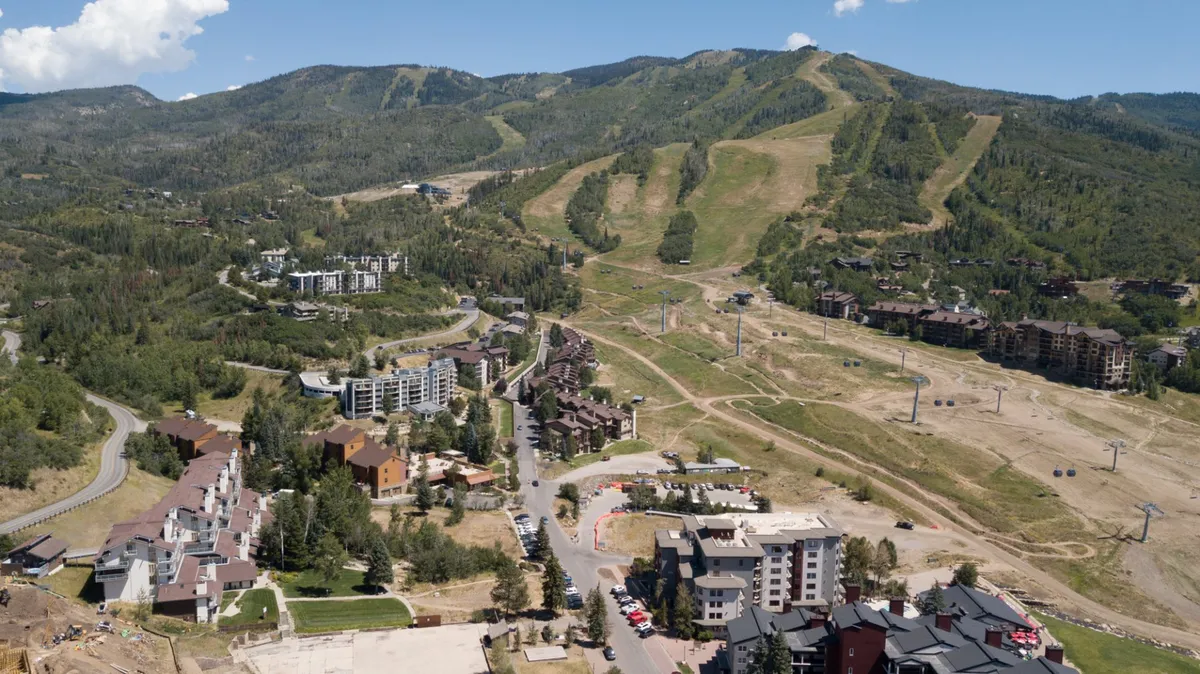 A sweeping exterior view focuses on the snow-covered mountain slopes and ski lifts, with residential resort buildings nestled at the base. A winding road and nearby trails are visible, set beneath a serene sky, highlighting both the adventure and tranquility of the locale.