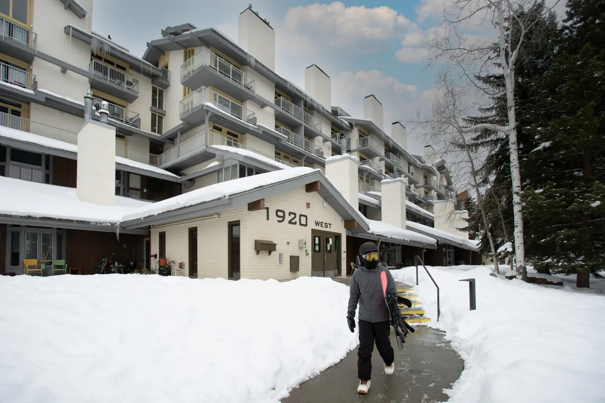 A snow-dusted exterior shows the building facade with a snowy pathway leading toward it. A person carrying a snowboard is visible, suggesting direct access to the slopes. The scene captures vibrant mountain life with people walking and socializing near sunset, while balconies overlook the snow and mountain views.