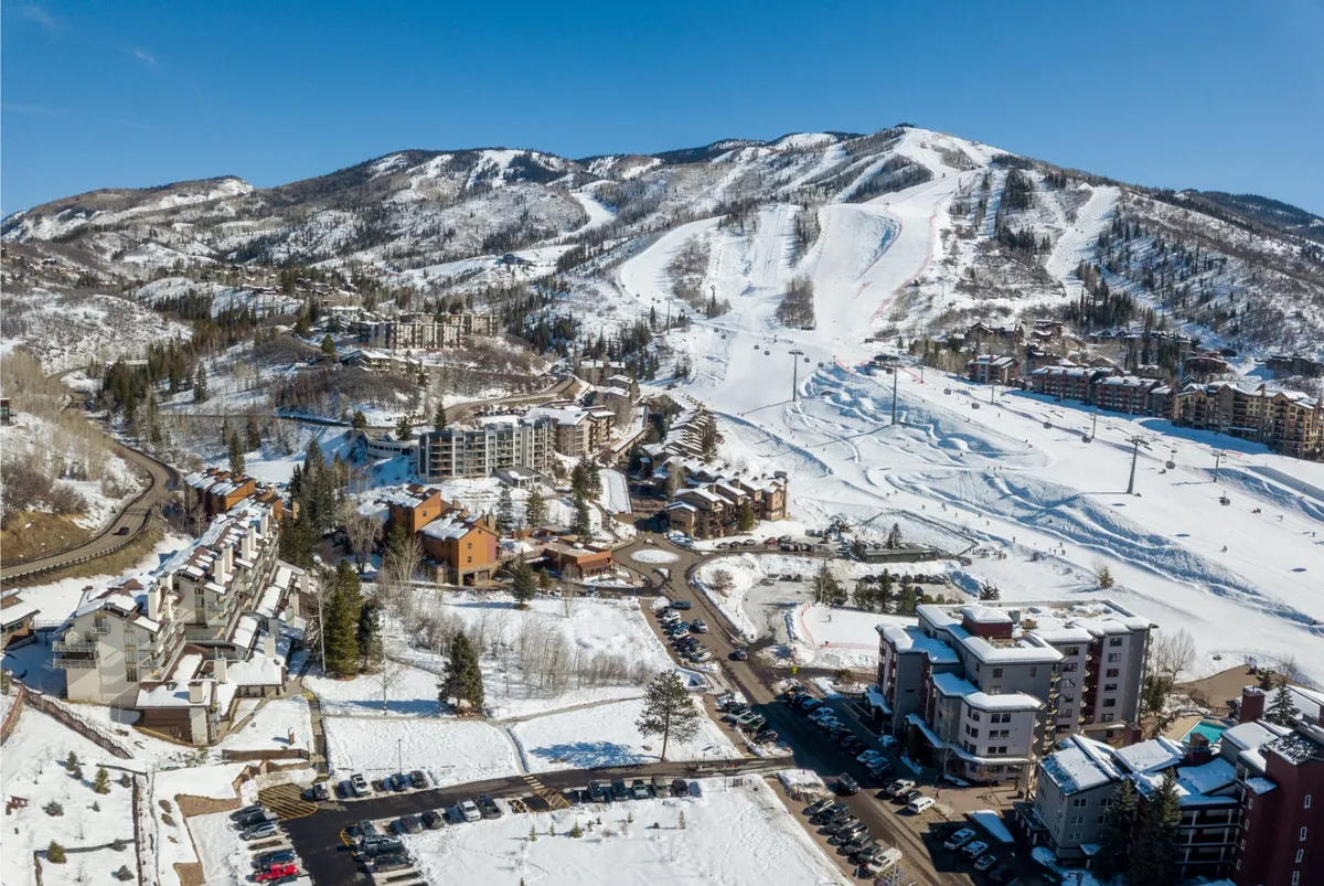 A panoramic exterior shot highlighting snow-blanketed ski slopes, resort buildings, and a spacious parking area, with sunrise glinting over the mountains. The scene suggests both tranquility and excitement, suitable for various mountain activities.