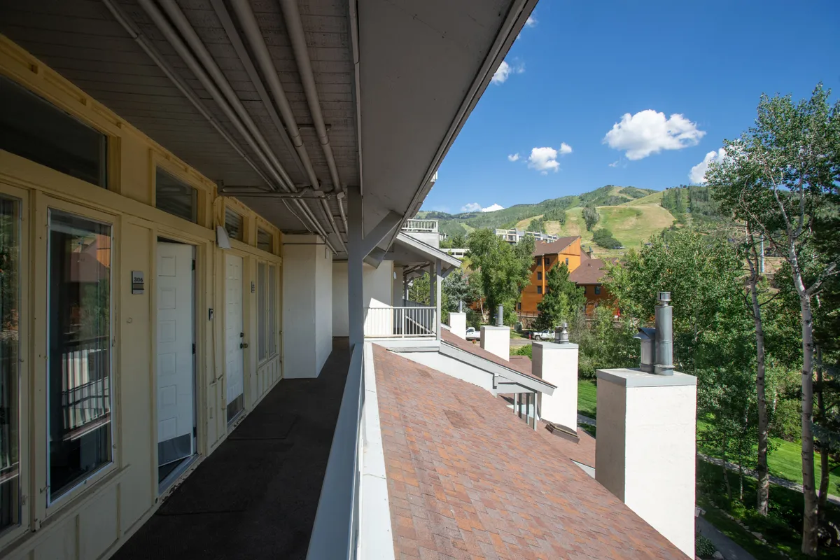 A peaceful balcony scene overlooking picturesque mountain peaks, colorful resort buildings, and lush greenery. The vantage point immerses viewers in the alpine setting and hints at the proximity of local attractions and community events.