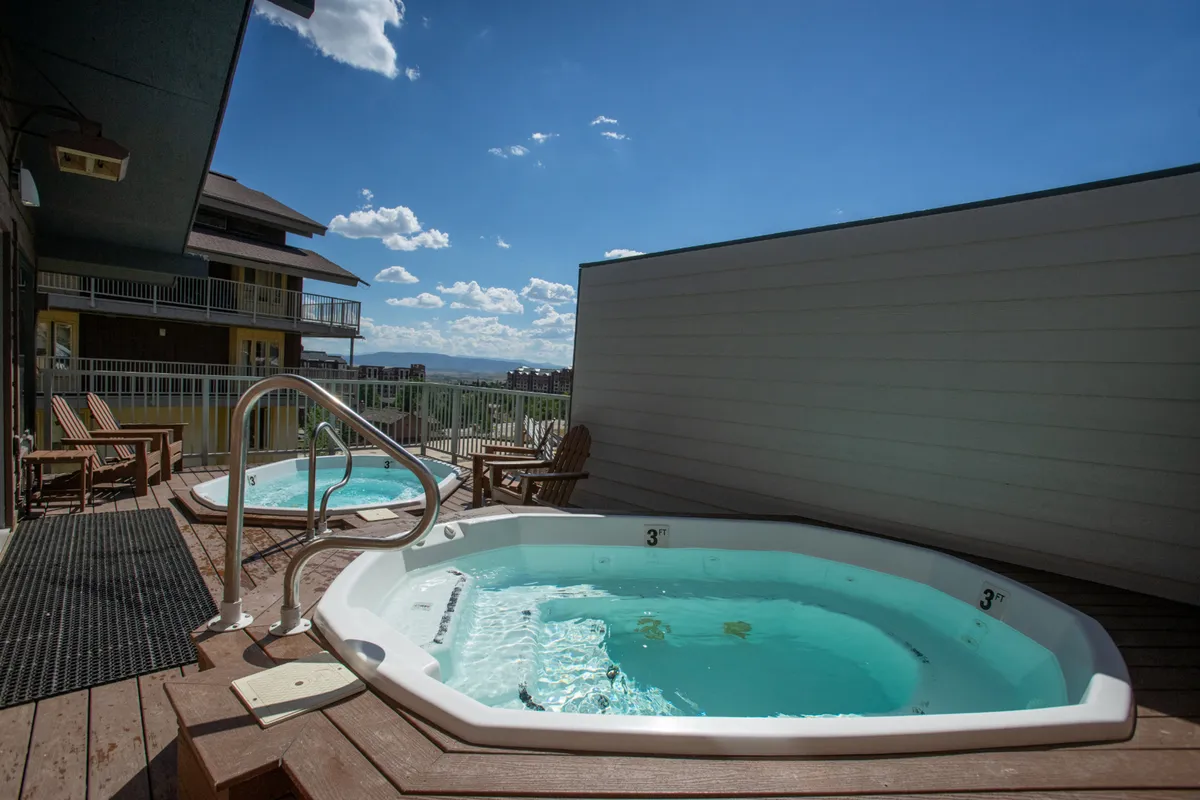The image displays a tranquil outdoor setting with inviting hot tubs on a wooden deck, surrounded by lounge chairs and offering a scenic mountain view. The space suggests relaxation after outdoor adventures, with both daytime and nighttime appeal for stargazing or enjoying a morning coffee.