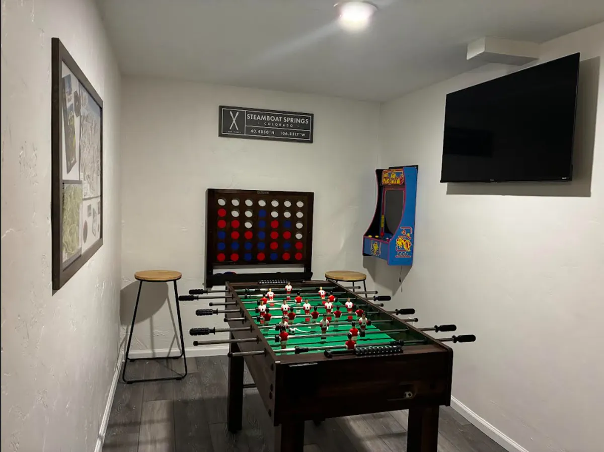 A vibrant game room featuring a foosball table in the foreground, a classic arcade game along the wall, bar stools for spectators, and art that gives the space a playful atmosphere. The setup invites friendly competition and social gatherings.