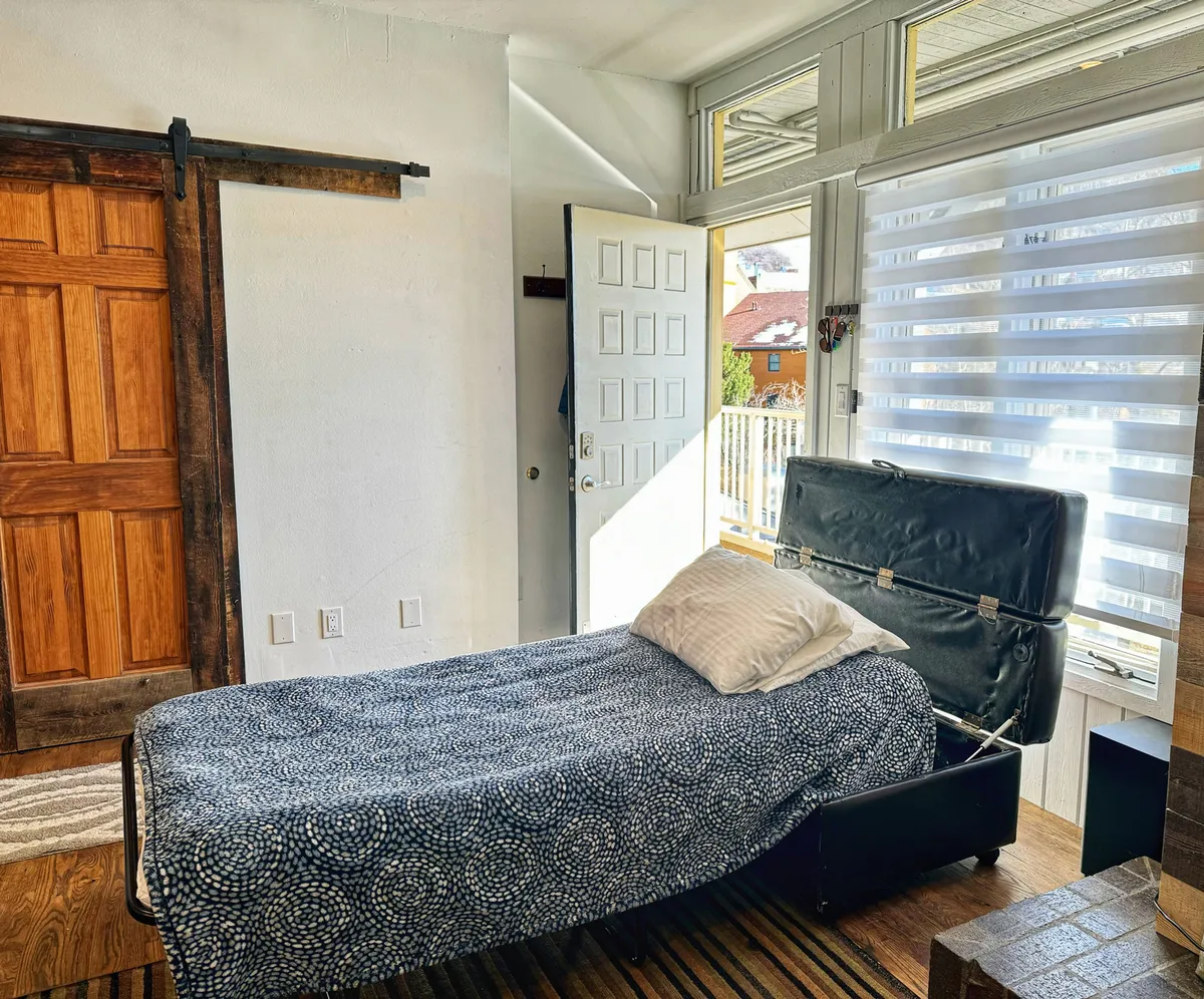 A serene lower-level bedroom is shown with a plush bed, an open door, a sliding barn door, and a bright window with blinds. The space feels comfortable and private, suggesting a sanctuary perfect for couples or anyone valuing seclusion and relaxation.